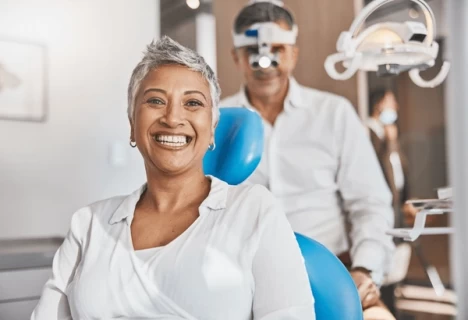 "Smiling woman sitting in a dental chair with a dentist in the background wearing magnifying loupes, in a modern dental office."