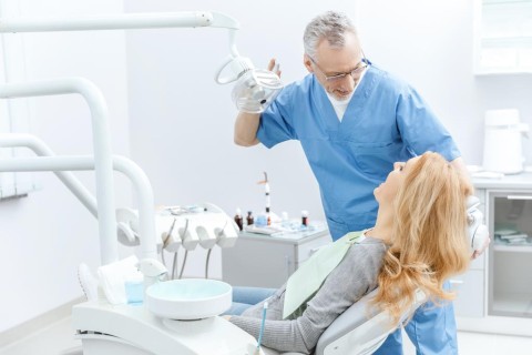 A male dentist in blue scrubs adjusts a dental light while interacting with a female patient in a modern clinic.