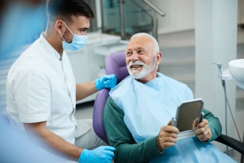 A senior man smiles at a dentist while holding a mirror in a dental clinic.