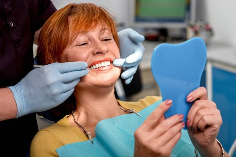 A smiling middle-aged woman with red hair is sitting in a dental chair, holding a blue tooth-shaped mirror while a dentist, wearing blue gloves, adjusts her dental prosthetics or veneers.