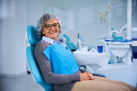 An elderly woman with gray hair and glasses is sitting in a dental chair, wearing a blue protective bib, smiling confidently in a modern dental office setting.